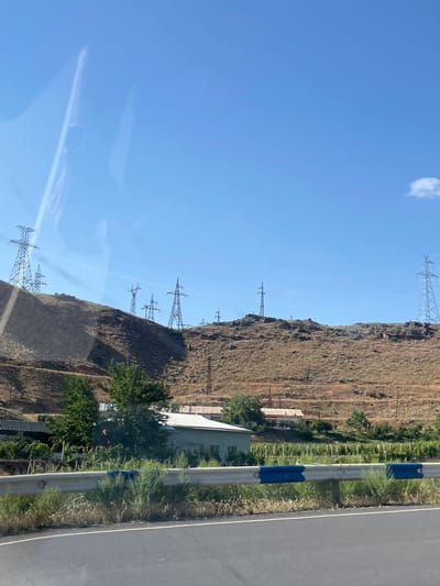 In the city of Gladzor, Armenia, a vast expanse of arid, brown hills dominates the landscape, punctuated by numerous tall electrical pylons stretching across the horizon. The sky is a clear, bright blue with a single small white cloud visible in the distance. In the foreground, a paved road runs horizontally, bordered by a metal guardrail with blue and white reflective markers. Beyond the guardrail, green foliage and what appear to be vineyards are situated on a lower slope. Several residential buildings are nestled amongst the vegetation, with pitched roofs and light-colored walls. The overall scene depicts a rural or semi-rural environment with significant electrical infrastructure.
