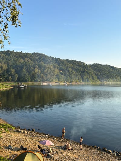 The image depicts a serene lakeside scene in Lipie, Poland. In the foreground, a rocky shore is dotted with camping equipment, including a green and orange tent, a pink umbrella, and various bags. Two individuals are visible near the water's edge: one person, wearing black shorts, stands and appears to be fishing, while another person in a striped top and dark shorts stands nearby. Further along the shore, another individual is seated, possibly preparing for fishing. A faint plume of smoke rises from the water between the two standing individuals. The lake itself is calm, reflecting the clear blue sky and the lush, tree-covered hillside that forms the backdrop. Several small boats are anchored in the distance along the shoreline. The overall atmosphere is one of tranquility and outdoor recreation.