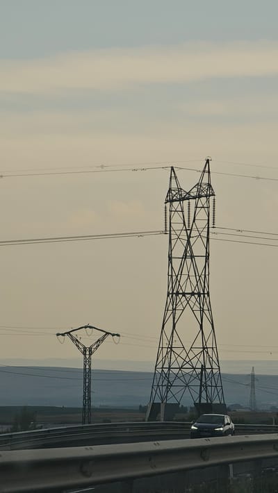 The image depicts a view from a highway overpass, looking towards a landscape with power lines and transmission towers under a hazy sky. A dark-colored car is driving on the highway in the foreground. In the middle ground, a large, dark metal transmission tower with a complex lattice structure is prominent. To its left, a smaller transmission tower with a diamond-shaped crossarm supports power lines. Further in the background, another transmission tower is visible. The landscape behind the towers consists of rolling hills or distant mountains, layered in muted blues and grays, suggesting a vast, possibly rural or semi-rural area. The sky is a pale, diffused color, indicative of overcast or foggy conditions. The overall impression is one of industrial infrastructure set against a somewhat muted natural environment, typical of a transitional zone between urban and rural areas. While the image does not explicitly show elements specific to Zaragoza, Spain, such industrial landscapes with transmission towers are common in the outskirts of many cities, including those surrounding Zaragoza, a city known for its plains and proximity to mountainous regions in Aragon.