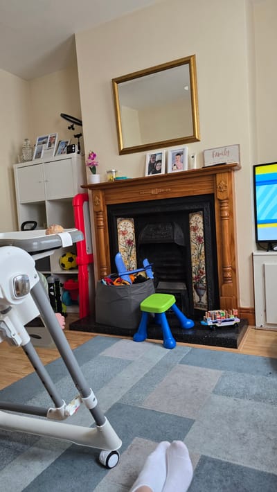 The image depicts an indoor scene, likely a living room in Kilkenny, Ireland. A traditional wooden fireplace with decorative tiles dominates the center of the frame. To the left of the fireplace, a gold-framed mirror hangs on a cream-colored wall, reflecting the room. Below the mirror and to the left of the fireplace, a white shelving unit is visible, adorned with framed photographs and a small pink orchid.

In front of the fireplace, a grey fabric toy bin is overflowing with children's toys, including a blue toy airplane and a green stool with blue legs. A toy car made of wooden blocks is placed on the black granite hearth. Adjacent to the fireplace, a red toy structure and a yellow soccer ball are scattered on the floor, suggesting active play. A baby high chair is positioned in the foreground, partially obscuring the view of the floor, which is covered by a patterned blue and grey rug. In the upper right corner, a pair of white socks are visible, indicating a person is present in the room, though only their feet are shown. Above the fireplace, a portion of a television screen displaying a colorful striped pattern is visible.

A sign with the word "Family" written in cursive is hanging on the wall above the mirror, with smaller text below it. The lighting suggests it is daytime, and no outdoor weather cues are apparent. The overall impression is a cozy, lived-in family space with a focus on children's activities.
