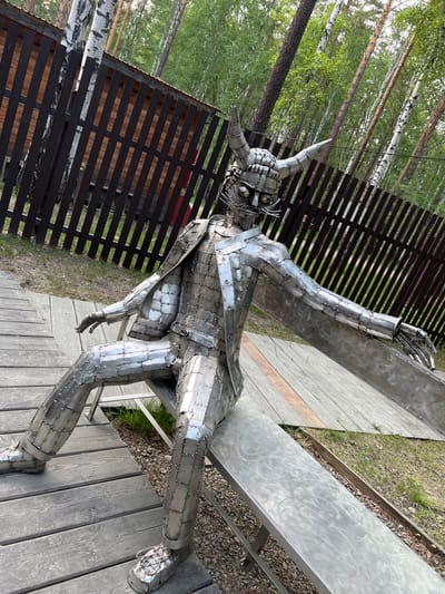 A full shot shows a metal sculpture of a seated figure with a horned helmet. The sculpture is made of numerous small, riveted metal plates, giving it a segmented appearance. The figure is wearing a suit jacket and trousers, and its arms are outstretched. The sculpture is sitting on a metal bench in an outdoor setting. The background consists of a wooden fence, a forest with birch trees, and a wooden deck. The location is the Taltsy Museum of Architecture and Ethnography in Tal'tsy, Russia.