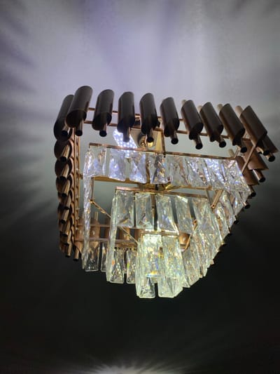 The scene is located in Abad Tukimachi, Uzbekistan. The image depicts an illuminated, multi-tiered chandelier viewed from a low angle, suspended against a light-colored ceiling that transitions to a dark background. The chandelier features a gold-toned metallic frame. Its uppermost section consists of numerous dark, semi-cylindrical, open-ended tubes arranged circumferentially and angled downward. Below this, multiple tiers display clear, rectangular, faceted crystal prisms, individually suspended. Active light sources within the chandelier emit bright light, which refracts through the crystal elements, creating spectral highlights and casting patterned shadows onto the ceiling and dark surroundings. No individuals or explicit actions are visible within the frame.