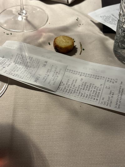 The image shows a restaurant table setting in Karneid, Italy. A half-eaten roasted potato sits beside a receipt. The receipt indicates a meal totaling €749.40, including items like Vodka Beluga, tonic, desserts, Tignanello wine, bottled water, polpettine (meatballs), ravioli, steak, and "ofenkarfoffel" (oven-baked potatoes) with grilled vegetables. The receipt has details of the establishment, including its name, address (Eggentaler Strasse 47), and tax information, alongside transaction details. A wine glass is also visible. The table has a textured tablecloth.