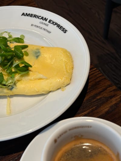 The image shows a close-up view of a breakfast setting on a wooden table, likely in the American Express Lounge at Stockholm-Arlanda Airport, Sweden. A white plate holds a folded omelet garnished with fresh green herbs. The plate is branded with "AMERICAN EXPRESS LOUNGE" and "BY PONTUS FRITHIOF" in black text. To the right, a white cup contains a dark espresso with a foamy crema. The cup also has faint gold text on its rim, reading "ARVID NORDQUIST". The background is blurred, showing the dark, grainy texture of the wooden table. There are no people visible in the image.