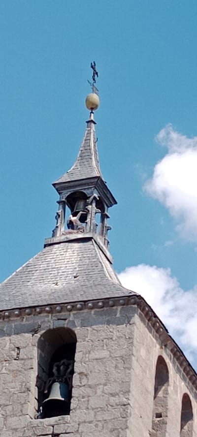 The image shows the upper portion of a stone church tower in Galapagar, Spain. The tower features a steeply pitched, grey-tiled roof that leads to a belfry with an open structure housing a bell. Above the belfry, a smaller spire is topped with a weather vane. The sky is clear and blue, with a few white clouds visible. The stonework of the tower is rough-hewn and appears to be ancient. There is no indication of any people or specific actions taking place within the frame. The focus is on the architectural elements of the tower and its surroundings under a bright sky.