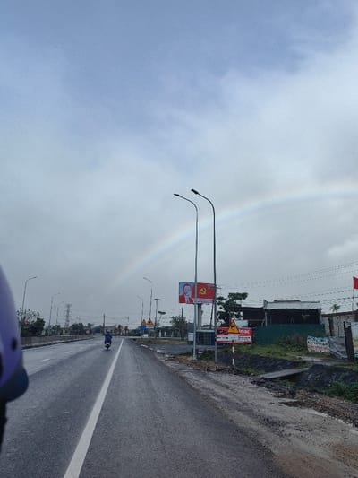 The image depicts a wet asphalt road extending into the distance under an overcast sky with a prominent rainbow arc. The scene is located in Hoành Sơn Ward, Vietnam.

In the foreground, the edge of a purple helmet is visible on the bottom left, indicating the perspective from a moving vehicle. The two-lane road features a solid white dividing line. Approximately midway down the road, a solitary individual is operating a dark-colored scooter or motorcycle, traveling away from the viewer.

Along the right side of the road, multiple elements are visible: a large billboard features a portrait of Ho Chi Minh beside the flag of the Communist Party of Vietnam (red with a yellow hammer and sickle). Two tall streetlights stand in front of this billboard. Further right, a series of smaller signs include a yellow triangular warning sign and a white rectangular sign displaying "IMAC" and a phone number. Beyond these signs, a drainage ditch with a small concrete bridge is visible, followed by vegetation and obscured residential or commercial structures. Overhead, numerous utility lines traverse the sky. A small red flag, likely a national or party flag, is visible on the far right.

On the left side of the road, a series of streetlights parallels the road, with distant power line towers and green foliage visible. The overall environment appears semi-urban or rural, characterized by recent rainfall evidenced by the wet road surface.
