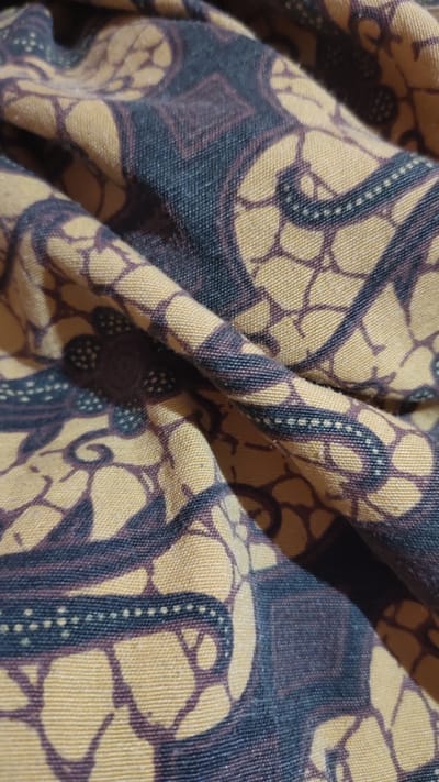 The image displays a close-up, high-angle shot of a textured fabric with a batik pattern. The pattern consists of interlocking dark purple and dark blue motifs on a yellow background. The fabric is creased and folded, creating shadows and highlights that emphasize its texture and depth. The overall impression is of a detailed textile, likely a sarong or similar garment, given the cultural context of batik in Indonesia. The image does not contain any identifiable landmarks or features that would allow for its specific location within Palangka Raya, Indonesia, to be determined.