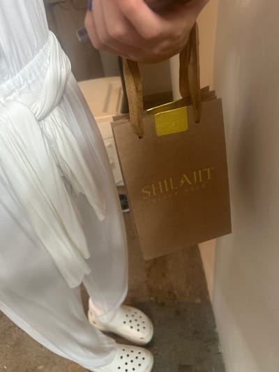 A person wearing a white jumpsuit and white clogs is holding a brown paper bag with gold handles. The bag has a gold label with the word "SHILAJIT" written on it in gold lettering. The label also reads "BLACK GOLD" in smaller lettering. The person is standing on a concrete floor. The coordinates provided are for a location in Pozos, Costa Rica, but the image does not show any specific features or details that would confirm that location.