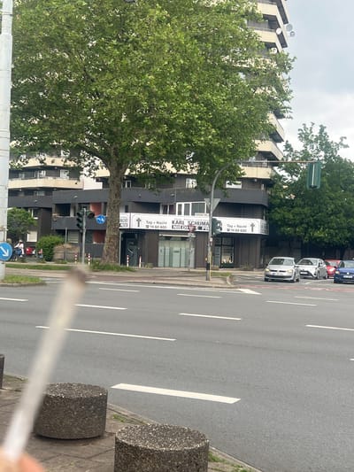 This media file captures a daytime street scene in Herne, Germany, under an overcast sky. The image is taken from street level, looking across a multi-lane road towards a row of buildings.
In the foreground, to the right, is a grey asphalt road with white lane markings. A concrete block is visible on the sidewalk beside the road. On the road, several cars are present, with a silver car prominently visible, followed by a red and a blue car.
Across the street, on the left, are several multi-story buildings, predominantly dark grey or black, with some lighter facades and window frames. Large, leafy green trees are scattered along the street, partially obscuring the buildings. One prominent tree stands in the mid-ground. The architecture of the upper parts of the buildings shows distinct, angled rooflines or structural elements.
Several signs and banners are attached to the buildings or poles. Visible text includes:
* "Sodinger Str." (Sodinger Street), indicating the street name.
* Banners for "KARL SCHUMACHER" with the text "Tag + Nacht" (Day + Night) and a phone number "0 23 23 / 14 60 600". Below "KARL SCHUMACHER", text also indicates "Feuer, Anonym und Erdbestattung" (Fire, Anonymous and Earth Burial), suggesting a funeral services business.
* Another sign with "Tag + Nacht" and the same phone number.
* The phrase "NIEDRIGE PREISE" (Low Prices) is also visible.
Traffic lights and street lamps are also part of the urban landscape. A blue circular sign indicating a shared path for pedestrians and cyclists is visible near the bottom left, pointing left. No people are clearly discernible, but the scene depicts a typical busy street environment.
