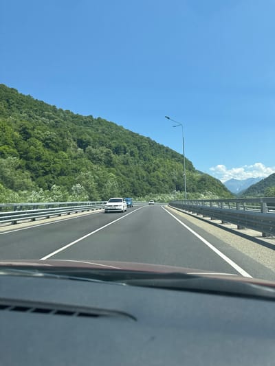 The image depicts a view from the perspective of a driver traveling on a highway. The highway is paved and has clear white lane markings. On the left side of the road, there is a metal guardrail. Beyond the guardrail, a steep, tree-covered mountain slopes upwards. The trees are lush and green, suggesting a warm season. The sky is a clear, bright blue with a few scattered white clouds visible in the distance, over a mountain range.

On the highway, several vehicles are visible. In the foreground, the dashboard and windshield of the car are visible, slightly obscuring the view ahead. Further down the road, a white car is traveling in the same direction, followed by a blue car. A lone white car is also seen further ahead on the left side of the highway. A street light stands tall on the right side of the road, casting no shadow due to the overhead sun.

The location is identified as Chvizhepse, Russia, a region known for its mountainous terrain and natural beauty, often characterized by such highways winding through valleys and alongside steep slopes. The overall atmosphere is serene and scenic, with the clear weather enhancing the visibility of the landscape.