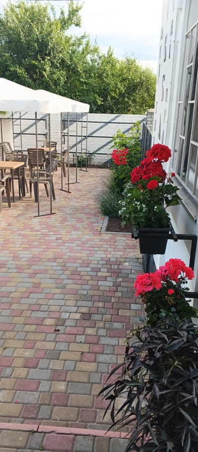 This image depicts an outdoor seating area with red flowers and dark green plants. The ground is paved with colorful interlocking bricks, a mix of red, pink, yellow, and grey. To the left, there are white umbrellas providing shade over several tables and chairs, made of dark plastic and wood. Behind the seating area, a metal fence with a white brick wall is visible. To the right, the exterior of a white building with windows and decorative black lettering can be seen. The scene is illuminated by natural daylight, suggesting it is daytime. The location is identified as Ploske, Ukraine.