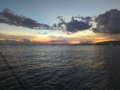 This image captures a marine scene at dusk or sunset in Port of Spain, Trinidad and Tobago. The sky exhibits a gradient of colors, transitioning from light blue at the zenith to orange and yellow hues near the horizon, interspersed with substantial dark grey cumulus clouds.
The lower two-thirds of the image comprise a wide expanse of dark, rippled water, reflecting the subdued light from above. Several vessels are present on the water: three sailboats with visible masts are distributed across the mid-distance, one on the far left and two towards the right. A single motorboat or similar small vessel, emitting a white light, is positioned on the far right midground. Two slender, dark vertical poles or stakes protrude from the water in the left foreground.
The distant horizon reveals a discernible coastline characterized by undulating hills or mountains. Numerous scattered artificial lights are visible along this landmass, indicating an urbanized area. No individuals are visible within the frame, and no specific actions or interactions are depicted.