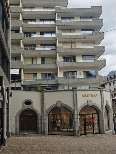 The image shows a street-level view of the Valentino store in St. Moritz, Switzerland. The store is located on the ground floor of a multi-story building. The building's facade is a mix of light-colored plaster and stone. The Valentino store has a large glass storefront with arched entrances. Inside the store, mannequins display clothing and handbags are arranged on shelves. Two people are visible inside the store.
To the left of the Valentino store, there is another building with a similar architectural style. Above the Valentino store, the multi-story building has multiple balconies with glass railings. The street in front of the store is paved with cobblestones. In the background, a mountain peak is visible. The sky is overcast.