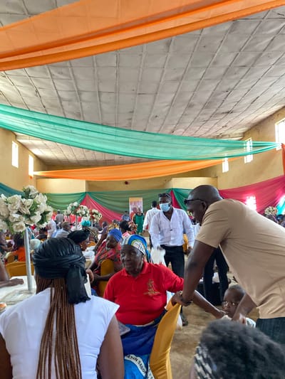 The image depicts an indoor gathering, likely a celebration or memorial service, within a building in Turu, Nigeria. The space is decorated with draped fabric in shades of orange, green, and pink, creating a festive atmosphere. The ceiling appears to be paneled with white squares.
In the foreground, a woman with braided hair wearing a white top is facing away from the camera. To her right, an older woman in a red shirt and a blue and white patterned headwrap is seated. Another man, wearing a tan shirt and sunglasses, is leaning over, seemingly interacting with a young child.
Several other individuals are visible in the background, seated at tables or standing. Some are wearing masks. The overall scene suggests a communal event with a focus on gathering and social interaction. The decorations and attire indicate a cultural context specific to the location.