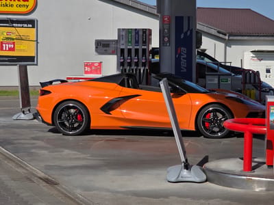This media file captures a bright, sunny day at a gas station in Rotmanka, Poland. The primary subject is an eye-catching orange sports car, possibly a Chevrolet Corvette C8, parked at a Verva fuel pump. The car's sleek design and vibrant color stand out against the neutral tones of the gas station building and pavement.
Other objects visible include multiple Verva fuel pumps with various fuel types labeled (98, 95, Diesel), a digital display on one pump showing "31.23 ZŁ 183.9 L," a vending machine or display for KitKat chocolates with a price tag of "3 1500," and a Pocztex parcel locker. Signs are present, including a large yellow and black advertisement in Polish for "NIISKIE CENY" (low prices) and grocery items like "Ptasie Mleczko" (Bird's Milk candy). Another sign prominently displays "VERVA" branding.
The scene appears to be during daylight hours, suggested by the strong shadows and bright illumination. The presence of the sports car at the fuel pumps indicates the activity of refueling. The overall impression is a modern, functional gas station setting with a touch of luxury from the sports car. The weather seems pleasant, with clear skies contributing to the bright atmosphere. There are no people directly visible in the image, focusing the viewer's attention on the car and the gas station environment.