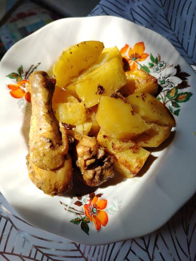 A close-up shot features a white, floral-patterned plate filled with roasted chicken and potatoes. The chicken leg is prominently placed on the left side of the plate, appearing golden brown and seasoned. Alongside the chicken are several wedges of roasted potatoes, also golden and slightly browned, some with visible specks of seasoning or herbs. The plate is set on a patterned tablecloth. The lighting suggests natural light, casting shadows across the food and plate. The image is taken in an indoor setting, likely a home, in Жернов ауылдық округі, Kazakhstan.