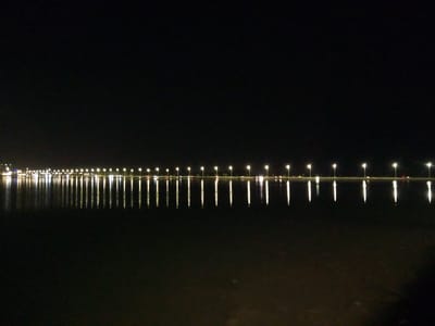 The image depicts a long, straight line of illuminated lampposts stretching across the horizon at night. The lampposts are spaced evenly apart and cast reflections on the still body of water below. The water is dark and reflects the lights, creating a symmetrical pattern. The sky is completely black, indicating nighttime. The scene appears to be a waterfront area or a road alongside a large body of water. This location is likely the Al Khobar Corniche in Al Khobar, Saudi Arabia, a well-known waterfront promenade lined with lampposts and offering views of the sea. There are no people or active events visible in the image, presenting a serene and static nocturnal landscape.