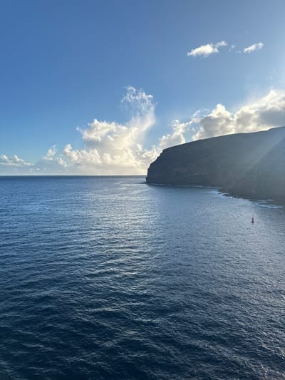 The image displays a coastal panorama near San Sebastián de la Gomera, Spain. The scene is dominated by a deep blue ocean that fills the left two-thirds of the frame, characterized by small ripples and a reflective band of sunlight extending from the horizon towards the viewer. On the right, a dark, steep, and rugged cliff face rises from the water, exhibiting distinct horizontal formations or striations across its surface. The base of the cliff meets the ocean with a rocky shoreline. In the lower right quadrant, a single red conical buoy with a white top floats on the water, positioned near the cliff base. The sky above is clear blue with scattered white cumulus clouds, particularly dense over the horizon on the left and above the center. Sunlight, partially obscured by clouds, casts bright rays across the upper portion of the image and illuminates the clouds from behind, suggesting an early morning or late afternoon setting. No individuals, vessels (other than the buoy), or direct actions are visible.