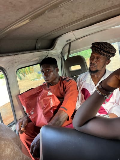 Two men are seated inside a vehicle, possibly a bus or van, in Okwe, Nigeria. The man in the foreground, on the left, is wearing an orange garment and holding a red bag with Chinese writing. He has dark skin and short black hair. He is looking forward, and his hands are clasped. To his right sits another man wearing a white shirt with pink accents and a patterned cap. He has a darker complexion and a beard, and he is looking down and to the side. The interior of the vehicle appears worn and functional. Through the window, a dusty, outdoor environment with some vegetation and structures is visible. The lighting suggests it is daytime. The focus is on the two men and their immediate surroundings within the vehicle.