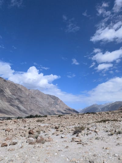 The image depicts a mountainous, arid landscape with a rocky foreground. The sky is a clear blue, dotted with white cumulus clouds. In the background, large, rugged mountains dominate the horizon, their slopes displaying various shades of brown and gray. Some of the peaks in the distance appear to be snow-capped. The ground in the foreground is covered with numerous stones and pebbles of varying sizes, with sparse, dry vegetation growing between them. The overall impression is one of a remote and harsh natural environment. The scene is devoid of any human presence or activity. The location is identified as Sasoma, India.
