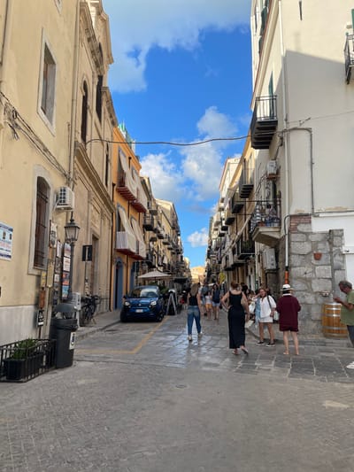 The image presents a narrow, paved street identified as Via Mandralisca in Cefalù, Italy, under a blue sky with scattered white clouds. The perspective looks generally southeast, with a sign on the left indicating "Piazza Duomo / Per Visitare / La Cattedrale Normanna."
The left side of the street is lined with a multi-story building featuring light-colored stucco, architectural moldings, and multiple windows, some with security bars. An exterior air conditioning unit is mounted near a lower window. A black lamp post with several affixed signs stands in the foreground, adjacent to a black public trash bin and a low black metal fence with plants. A black bicycle is parked against the building wall.
The right side of the street is bordered by a multi-story building with a base of rough-hewn stone blocks transitioning to light-colored stucco above. This building features multiple balconies with black wrought-iron railings, from one of which laundry is visible hanging. Several exterior air conditioning units are mounted on its facade. A large wooden barrel is positioned against the building's stone base.
A dark blue Fiat Panda automobile is parked on the left side of the street, facing away from the viewer, with yellow painted lines on the pavement nearby. Further down the street, multiple A-frame signs and outdoor restaurant umbrellas are visible.
Approximately 15-20 individuals are present on the street. In the foreground, two women walk away from the camera. In the mid-ground on the right, a group of 5-6 individuals are standing, including a woman in a white hat and dark red clothing, another woman who is barefoot, and a man in a green shirt examining a mobile phone. Additional individuals are visible walking or standing further into the background. The scene is illuminated by bright daylight, casting visible shadows.