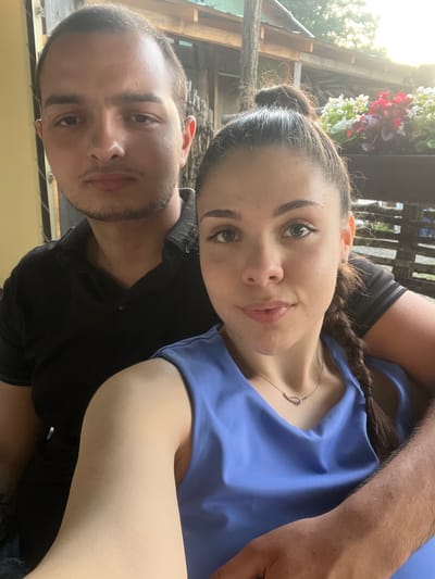 In this close-up selfie, a young couple is captured outdoors, leaning into each other. The woman, in the foreground, tilts her head towards the camera, her dark hair styled in a braid that rests on her shoulder. She wears a blue top and a delicate silver necklace. Her eyes are open, and she has a gentle smile. Behind her, a man's arm is visible, draped around her. His face, also visible, has short dark hair and a light beard. He is wearing a black shirt.

The setting appears to be a rustic outdoor space. In the background, sunlight streams through trees, casting a warm glow. Wooden structures and railings are visible, suggesting a porch or outdoor seating area. A cluster of red and white flowers adds a touch of color to the scene, enhancing the natural ambiance. The overall impression is one of a relaxed and intimate moment shared between the couple. The lighting suggests it is daytime, likely late afternoon due to the warm, angled sunlight. There is no visible text in the image.