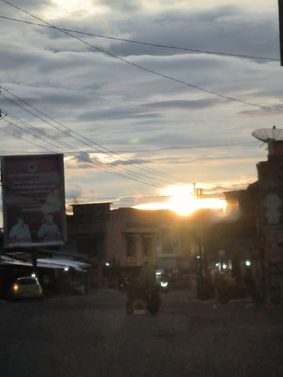 The image depicts a street scene in Jamurujung, Indonesia, during twilight. The sky is filled with dramatic, overcast clouds tinged with the warm glow of the setting sun, which is prominently visible on the right side of the frame, creating lens flare. Utility wires crisscross the sky, a common sight in urban environments.
In the foreground, a yellow car is parked on the left side of the road, its rear taillight illuminated. Further down the street, other vehicles, including what appears to be a motorcycle or scooter, are visible, some with their headlights or taillights on, indicating low light conditions. Buildings line the street, some with shopfronts or awnings. A large billboard is positioned on the left, displaying images of two individuals. On the right, part of a building with a satellite dish on its roof is visible. The overall impression is of a busy street as dusk settles.