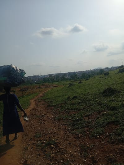 A person, viewed from the side and from the knees down, walks along a dirt path on a grassy, hilly landscape. The person wears a blue dress and sandals, and carries a large, bundled item on their head. The bundle is dark and appears to be covered in a plastic bag. The person also carries a small, white object in their right hand. The path is composed of dirt and small stones, with sparse green vegetation growing on either side. The background reveals a sprawling city with buildings and trees, possibly indicating a settlement in Tasha, Nigeria. The sky is a pale blue with scattered, light gray clouds. The overall scene depicts a person engaged in carrying goods through a rural or semi-urban environment.