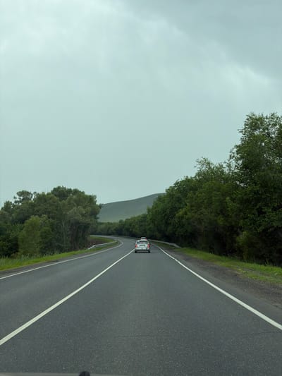A vehicle, a silver sedan with a visible license plate, is traveling on a two-lane asphalt road in Верхнеозёрное, Russia. The road is marked with a solid white line on the left and a broken white line on the right, indicating a clear lane separation. The car is in the right lane, heading away from the viewer. The environment consists of lush green trees lining both sides of the road, with a gentle hill in the background. The sky is overcast and grey, suggesting a cloudy or possibly rainy day. The overall scene depicts a scenic drive through a rural or natural landscape.