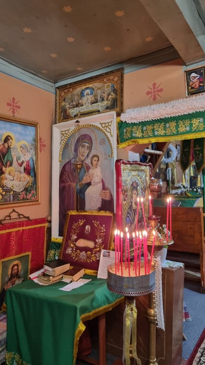 This image depicts the interior of an Eastern Orthodox church in Vyshka, Ukraine, possibly during a religious service or a moment of prayer. The scene is rich with religious iconography and ceremonial objects.
Dominating the foreground are several icons, including a large, framed painting of the Virgin Mary holding the Christ Child. To its right, a smaller icon shows the same subject, with numerous red candles burning in front of it, their flames casting a warm glow. A metallic candelabra holds many of these candles. Another framed icon to the left depicts a biblical scene, possibly the Last Supper. Below these, another icon shows the Holy Family.
The church's walls are painted a soft peach color and adorned with decorative star patterns and a floral motif. Green and red liturgical cloths, edged with gold trim, are draped over surfaces and along the altar area, indicating a sacred space. A wooden lectern or table is visible in the background, with books and papers placed upon it. The overall atmosphere is one of reverence and devotion. The lighting suggests it is daytime, but the exact time is not discernible. The presence of icons, candles, and liturgical vestments are clear indicators of the religious setting. There is no discernible text in the image.