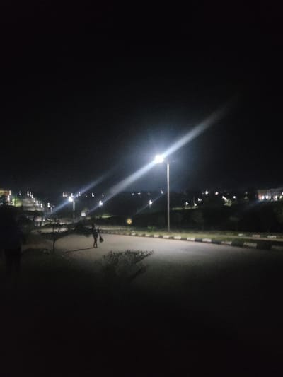 This image depicts a nighttime scene on a paved road in Gaba, Nigeria. The road, featuring black and white striped curbs on the right, recedes into the distance under the illumination of multiple streetlights. A prominent streetlight on the right emits a strong diagonal lens flare. One individual is visible walking on the road, slightly left of center, carrying a dark object in their left hand. A second, blurred figure is partially discernible on the far left foreground. The surrounding environment consists of dark terrain and low vegetation bordering the road, with distant artificial lights visible in the background.