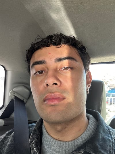 The image depicts a male adult seated inside a vehicle in Neuilly-Plaisance, France. The subject has a medium complexion, dark curly hair, and brown eyes, looking directly forward. A small red mark is visible on his forehead, and a mole is present below his right eye. He wears a small silver hoop earring in his right ear. His attire consists of a grey knitted high-neck sweater under a dark denim jacket. A dark blue seatbelt diagonally crosses his torso from his left shoulder. The vehicle's interior features a grey upholstered headliner and a dark headrest behind the subject's right side. Strong sunlight enters from the right, casting a distinct diagonal shadow across the subject's face, notably affecting his left eye and cheek. The exterior environment visible through the vehicle windows is largely indistinct, showing bright, overexposed areas to the left and faint details of a white railing and blue objects to the right.