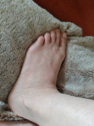 The image shows a close-up view of a human foot, resting on a furry, light brown surface. The foot is pale and appears to be relaxed. The toes are visible and the skin on the top and side of the foot shows natural texture and some wrinkles. The background is a darker, reddish-brown surface. There is no discernible connection to the city of Sumgayit, Azerbaijan.