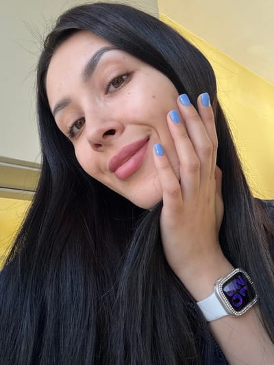 A female individual is depicted in a close-up portrait, presumably within an indoor setting in Molodezhnyi, Russia. She possesses long, dark, straight hair. Her complexion is fair, and she has light brown eyes with visible eyelashes and defined eyebrows. Her lips are adorned with light pink matte lipstick, and she exhibits a slight smile, showing subtle dimples. Her right hand is positioned against her right cheek, fingers slightly splayed, revealing short, square-shaped fingernails painted a light periwinkle blue. On her left wrist, she wears a smartwatch featuring a white band and a silver-toned case embellished with a double row of small, clear, brilliant-cut stones around the bezel. The watch face displays digital characters in a purple hue. A dark garment, likely a top or dress, is partially visible on her left side. The background is indistinct, consisting of a solid light yellow or cream color on the right, transitioning to a lighter tone on the left with a faint horizontal line.