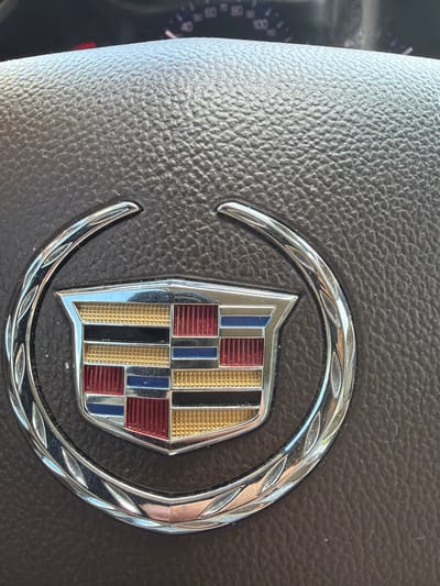 The image shows a close-up of a Cadillac emblem on a car's steering wheel. The emblem is silver with a crest in the center featuring red, blue, black, and gold colors. The steering wheel is made of dark, textured material. In the background, a blurred car dashboard with a speedometer is visible. The scene appears to be indoors or in a shaded area, as indicated by the lighting and the lack of strong shadows. The location is not specified as being in Grapevine, United States of America.