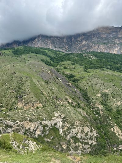 The image depicts a steep, verdant mountainside in Verkhnyaya Balkariya, Russia. The left side of the frame is dominated by a sheer, rocky cliff face partially obscured by thick, grey clouds and mist, suggesting a cool, possibly damp, and overcast day. The rock strata of the cliff are visible, showing a rugged, layered texture.

To the right of the cliff, the mountain slopes are covered in lush green grass and scattered trees, creating a stark contrast between the barren rock and the vibrant vegetation. The terrain is uneven, with visible rocks and patches of darker, steeper slopes that appear to be scree or eroded areas. The sunlight, though diffused by the clouds, illuminates the grassy slopes, highlighting the textures of the grass and the subtle variations in the terrain.

In the far right of the image, a patch of bright green grass suggests the edge of the mountainous area, possibly leading to a lower elevation or a different landscape. The overall impression is one of a wild, remote, and dramatic natural landscape. The weather conditions, with the encroaching mist and clouds, add a sense of mystery and vastness to the scene. There are no people or man-made objects visible, emphasizing the untouched nature of this mountain environment. The time of day appears to be daytime, given the natural light, but the overcast conditions prevent clear identification of the sun's position.