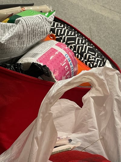 The image presents an overhead, close-up view into an open, reddish-orange bag or piece of luggage resting on a light grey floor. The bag is filled with various items. Prominently featured is a pink-wrapped tissue roll with "TISSUE ROLL" and "HUMANITARIANDARD" visible on its label. Adjacent to it are two packaged food items: a green and white packet labeled "NAAMKEEN" displaying nutritional information and allergen advice, and another white packet showing similar nutritional data. Behind these items, a black and white geometric patterned fabric item, possibly a pouch, is present, alongside an orange zipper pull tab. A crumpled white plastic shopping bag occupies the foreground, partially obscuring its contents, and faint non-English text is visible through its material. A black plastic bag is partially discernible beneath the pink tissue roll. No individuals are present. The scene is located in Seeb Province, Oman.