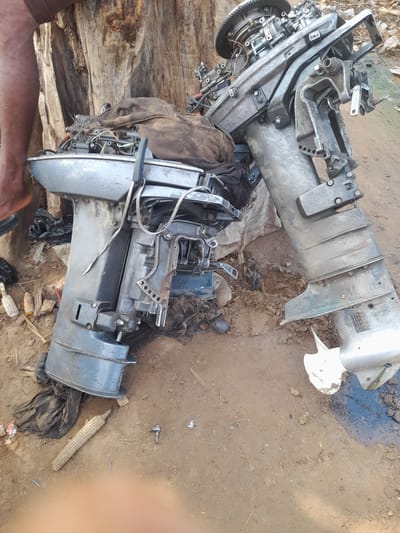 In Nemnei, Nigeria, two outboard motors are positioned on the ground. The motors are disassembled, with one motor positioned upright and the other lying on its side. One motor is gray and appears to be made of aluminum. The other motor is also gray but is darker in color and appears to be made of steel. Both motors are covered in dirt and debris. The area surrounding the motors is a dirt path, with some debris scattered around. A tree trunk is visible in the background on the left side of the image. A person's leg is visible on the left side of the image.