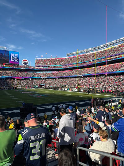 A football stadium packed with fans on a sunny day. The field is visible, with painted lines and logos. A stadium billboard is visible, promoting "Super Bowl LX". Spectators wearing jerseys and casual clothing occupy the stadium seating. A few fans wear blue and green, suggesting support for the Seattle Seahawks. The sky is clear blue.