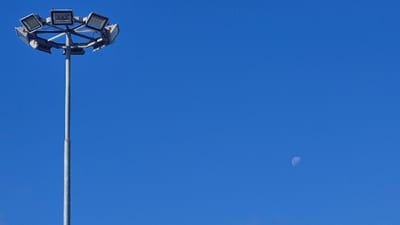 The image displays a clear daytime sky featuring a tall light pole and the moon. On the left side of the frame, a slender, grey metallic light pole stands vertically, extending towards the top. At its apex, a circular array holds approximately eight square-shaped floodlights, likely LED fixtures, positioned to project light outwards. These lights are off, consistent with the bright daytime conditions.

The sky is a vivid, deep blue, completely devoid of clouds, indicating excellent weather. In the upper right portion of the sky, a pale, half-crescent moon is distinctly visible against the blue background, highlighting that the photograph was taken during daylight hours.

There are no people present, and no specific activities are taking place. The scene is static, focusing on the infrastructure and the natural sky. There is no visible text in the image. The setting suggests an outdoor area, possibly a public space, industrial zone, or sports facility in Hualpén, Chile, where such high-mast lighting is commonly used.