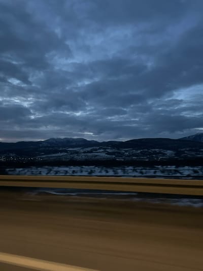 A view from inside a vehicle. Dark, turbulent clouds fill the sky, suggesting a storm. A mountain range is visible in the distance, with a few lights indicating a settlement at its base. A window frame is prominently featured, with motion blur on the right side, implying movement. The overall tone is somber and evokes a sense of travel through a potentially wild landscape.