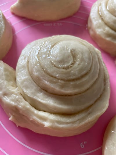 A close-up, overhead shot captures a batch of raw, rolled dough arranged on a pink silicone baking mat. The dough is shaped into spiral forms, similar to cinnamon rolls. The center of the main dough spiral is glistening with oil, revealing concentric layers of dough. Other similar dough shapes are partially visible in the background and on the right side of the frame. The baking mat has white markings, including a "6"" measurement indicator at the bottom right. The scene is devoid of any human presence or elements that would indicate a specific city or location like Aropakkuzi, Russia.
