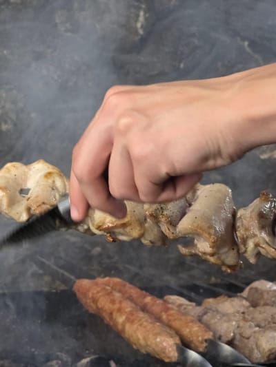 A close-up shot shows a hand manipulating meat on skewers over a dark, smoky background. The hand, with its pale skin, is positioned on the left side of the frame, fingers gently touching pieces of cooked meat threaded onto a metal skewer. The meat appears to be in various stages of cooking, with some pieces displaying a light brown char. The skewers themselves are visible, some holding chunks of meat and others holding what look like ground meat sausages. The smoky atmosphere suggests an outdoor cooking environment, possibly a barbecue or grill. The lighting is dim, indicating it might be evening or a shaded area. The overall scene evokes the preparation of a meal, likely a traditional dish involving grilled meat. The location context of Pidhorodne, Ukraine, hints at a possible cultural or culinary setting related to that region. There is no visible text in the image.
