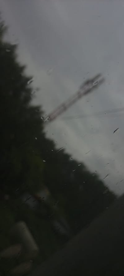 The image shows a view through a wet window, likely from inside a vehicle, looking outwards. The scene is overcast with a cloudy sky. In the foreground, there are water droplets on the glass, causing distortion. To the left, a dark, out-of-focus mass of foliage, possibly trees, dominates the lower portion of the frame. A blurred, reddish-brown, elongated object, possibly a construction crane or part of industrial equipment, is visible diagonally across the upper right quadrant of the image. The overall impression is one of a gloomy, possibly rainy day in an outdoor urban or semi-urban environment. The location in Kudymkar, Russia is not visually evident from the image itself.