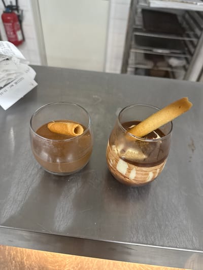This image shows two glasses containing desserts. The top glass has a dessert that appears to be chocolate mousse with swirls of white cream and a wafer cookie sticking out. The bottom glass contains a similar chocolate mousse dessert, also with a wafer cookie. Both glasses are on a metallic surface, possibly a counter in a kitchen or restaurant. In the background, there are shelves with items, and a red fire extinguisher is visible on the left. A receipt is partially visible in the foreground. The lighting suggests an indoor setting.
Visible text on the receipt reads "CUISINE mar. 19:44", indicating it is a kitchen receipt from Tuesday at 7:44 PM. The overall impression is a presentation of desserts in a professional food service environment.