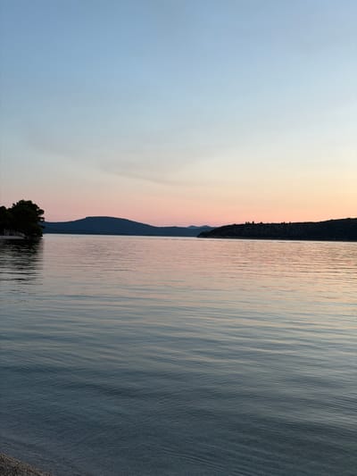 A serene coastal scene unfolds at sunset in Banići, Croatia. The sky transitions from a pale blue at the top to a soft pink and orange hue near the horizon, reflecting gently on the calm sea. In the distance, the dark silhouettes of rolling hills and islands create a dramatic backdrop. On the left side of the frame, a cluster of dark green trees juts out from a rocky shoreline, casting a shadow on the water. The foreground is occupied by the tranquil sea, its surface rippled by a gentle breeze, with a hint of a pebble beach visible at the very bottom edge. There are no people or animals present in the image.