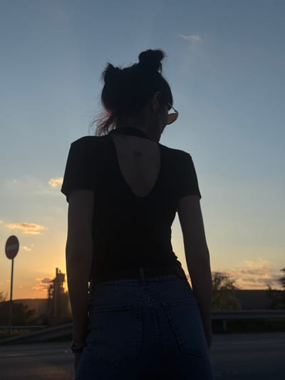 A woman stands silhouetted against a sunset sky. She has her hair in a bun, wears sunglasses, a dark top with an open back, and denim jeans. The background reveals a roadside with infrastructure, including a no entry road sign and foliage, suggestive of a location in or near the city of Kipra, Bulgaria.