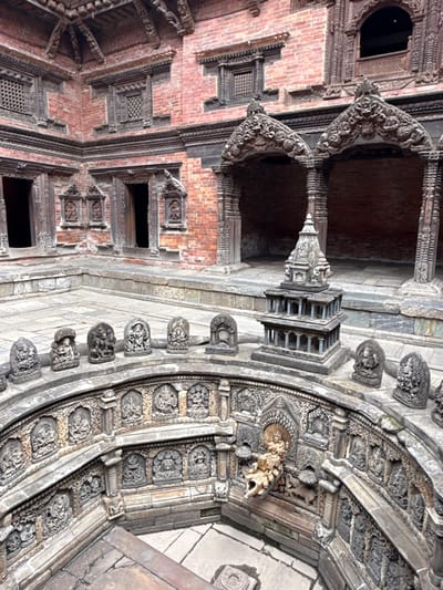 This image shows an architectural detail within a building in the city of Lalitpur, Nepal. The architectural elements include ornate carvings, brickwork, and multiple archways. A tower-like structure is visible in the center of the composition. A curved stone wall is adorned with a row of carved figures and a lion's head. Various windows and carved panels are present on the brick walls. The structure appears to be historical and possibly part of a temple or palace complex. The lighting is natural and somewhat muted, giving the stone and brick a textured appearance.