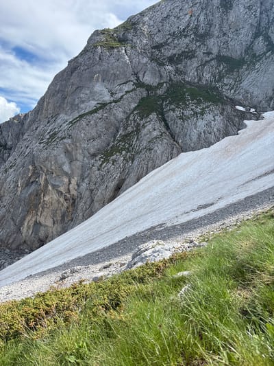 The image depicts a mountainous landscape in the vicinity of Pašina voda, Montenegro. A large, steep, grey rocky mountain dominates the upper left and center of the frame. Patches of green vegetation are visible on the mountain's surface, along with areas of snow. A large expanse of snow covers the slope in the lower right portion of the image, appearing to be melting or compacted. The foreground features green grass and low-lying, dense green shrubbery. The sky above is partly cloudy with streaks of white clouds against a blue background. The scene is natural and appears to be an outdoor, rugged terrain. No people or man-made structures are visible.