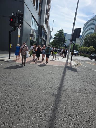 This media file depicts a street scene in Cardiff, United Kingdom. Several people are walking along a pavement next to a road. The time of day appears to be daytime, with shadows indicating bright sunlight. The weather seems to be pleasant.
In the foreground, a traffic light shows a red signal, suggesting that pedestrians may be waiting to cross. To the left of the traffic light, a modern building with large windows dominates the background. Further down the street, more people are visible, some walking in groups. One person is wearing a bright floral shirt, and another is in a blue dress. A person in a yellow high-visibility vest is also present.
The pavement is made of red and grey paving stones, and the road is asphalt. A shadow of a tall pole or light fixture stretches across the road. In the background, there are trees and more buildings, including one with a distinct blue facade. A car is visible on the road.
The overall atmosphere is that of a normal day in a city, with people going about their business. There is no indication of any specific event taking place, other than people using the street and crossing. Some visible text includes a "15" on a pole and what appears to be a business name "primark.com" on a shopping bag. The scene is one of urban activity under a clear sky.